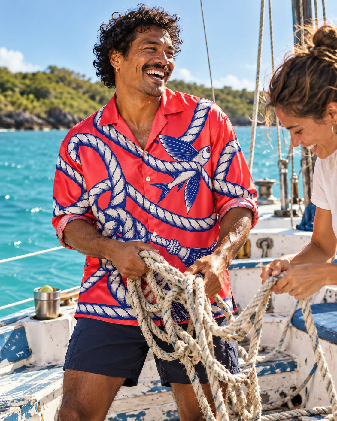 A curly-haired adult man laughing with a friend while untangling mooring rope aboard an old sailboat, wearing the coral Loose Ends silk shirt